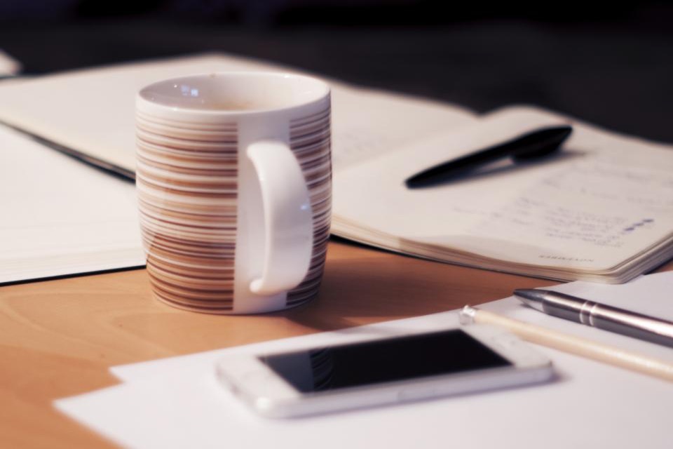 木の机に置かれたコーヒーカップと、開いたノートとペン、その手前のスマートフォン